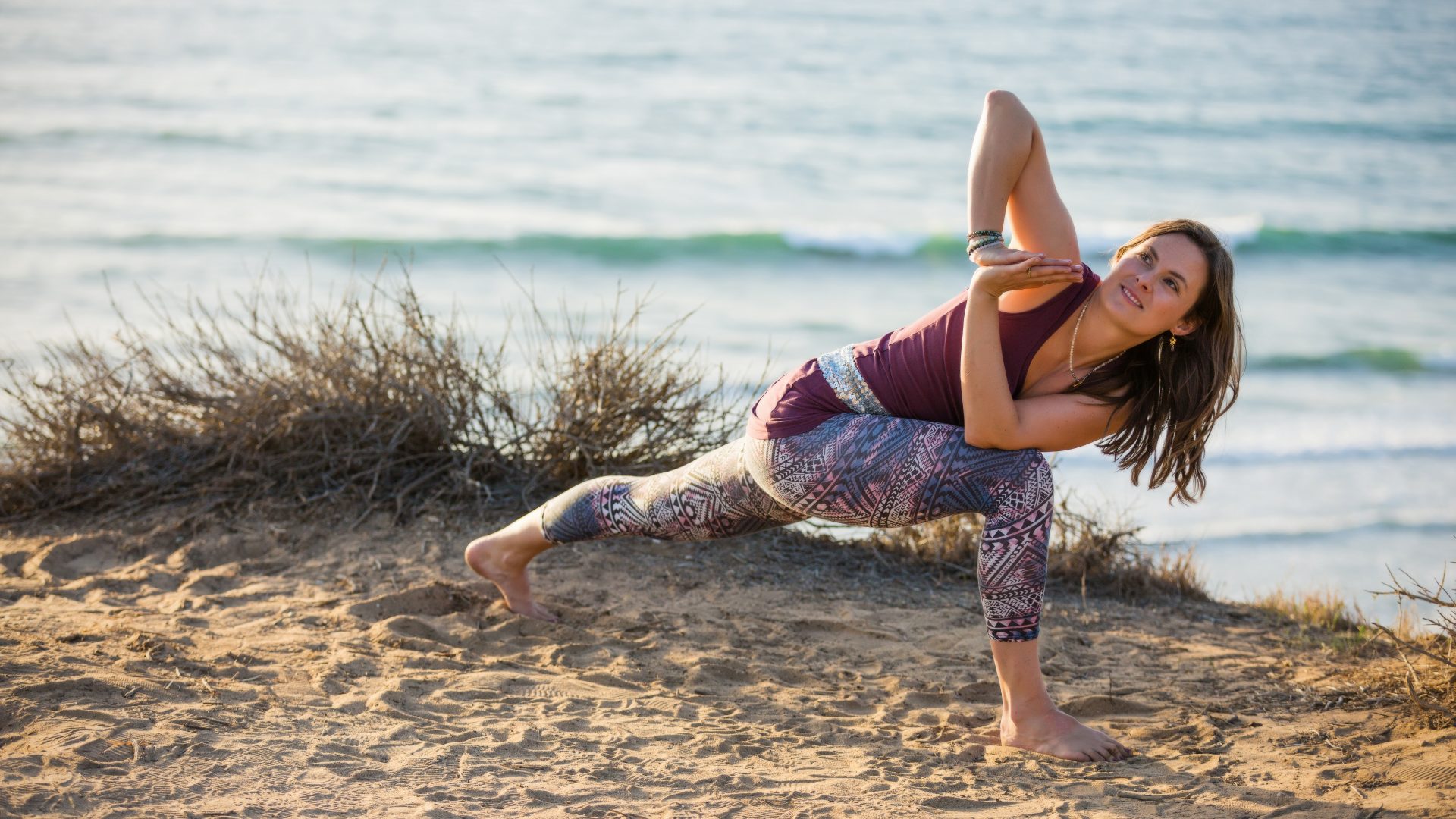 Kvinde på en strand, som poserer i en yogastilling. Iklædt blåt og lilla tøj med en masse mønstre på hendes tights. I yogastillingen ser hun op mod himlen og smiler.